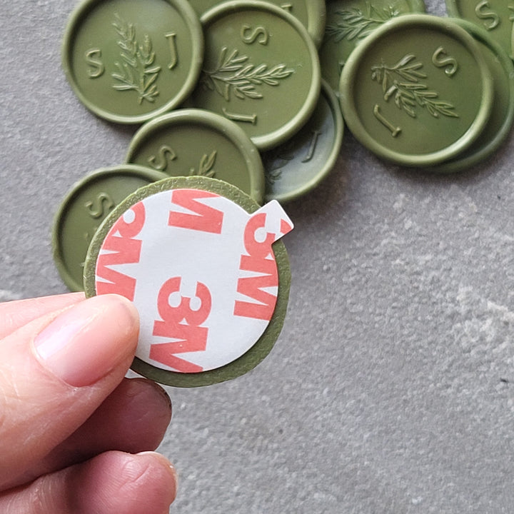 Close-up of a green 'Willow' monogram wax seal with 3M peel-and-stick adhesive backing, perfect for personalized stationery.