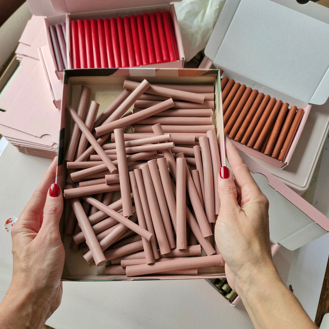 Hands holding a box of custom-colored wax sticks in a light pink shade, surrounded by boxes of red and brown wax sticks. Perfect for precision color matching.