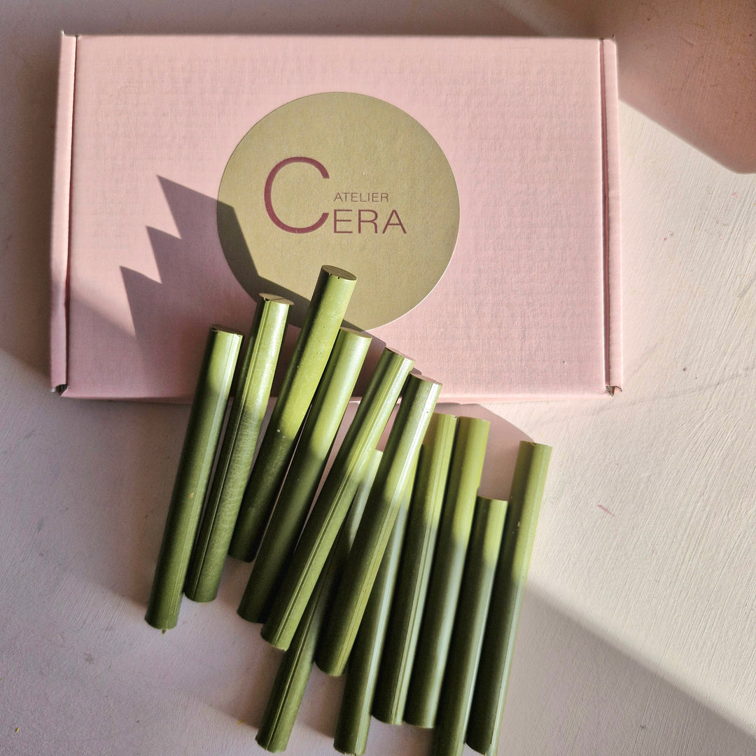 250 olive green 11mm sealing wax sticks arranged on a table with a pink Atelier Cera box in the background. Ideal for bulk sealing projects.