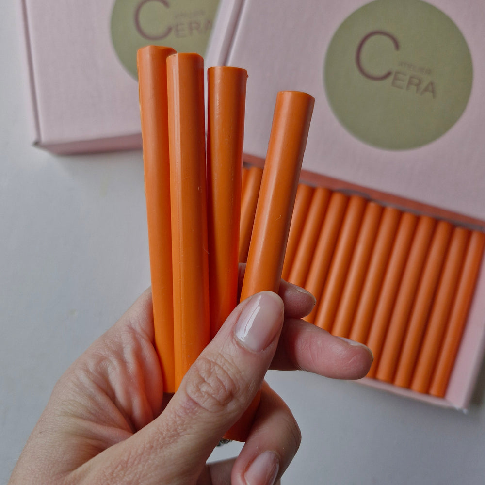 Close-up of 11mm tangerine sealing wax sticks held in hand, with a box of 30 in the background. Durable and easy to use.