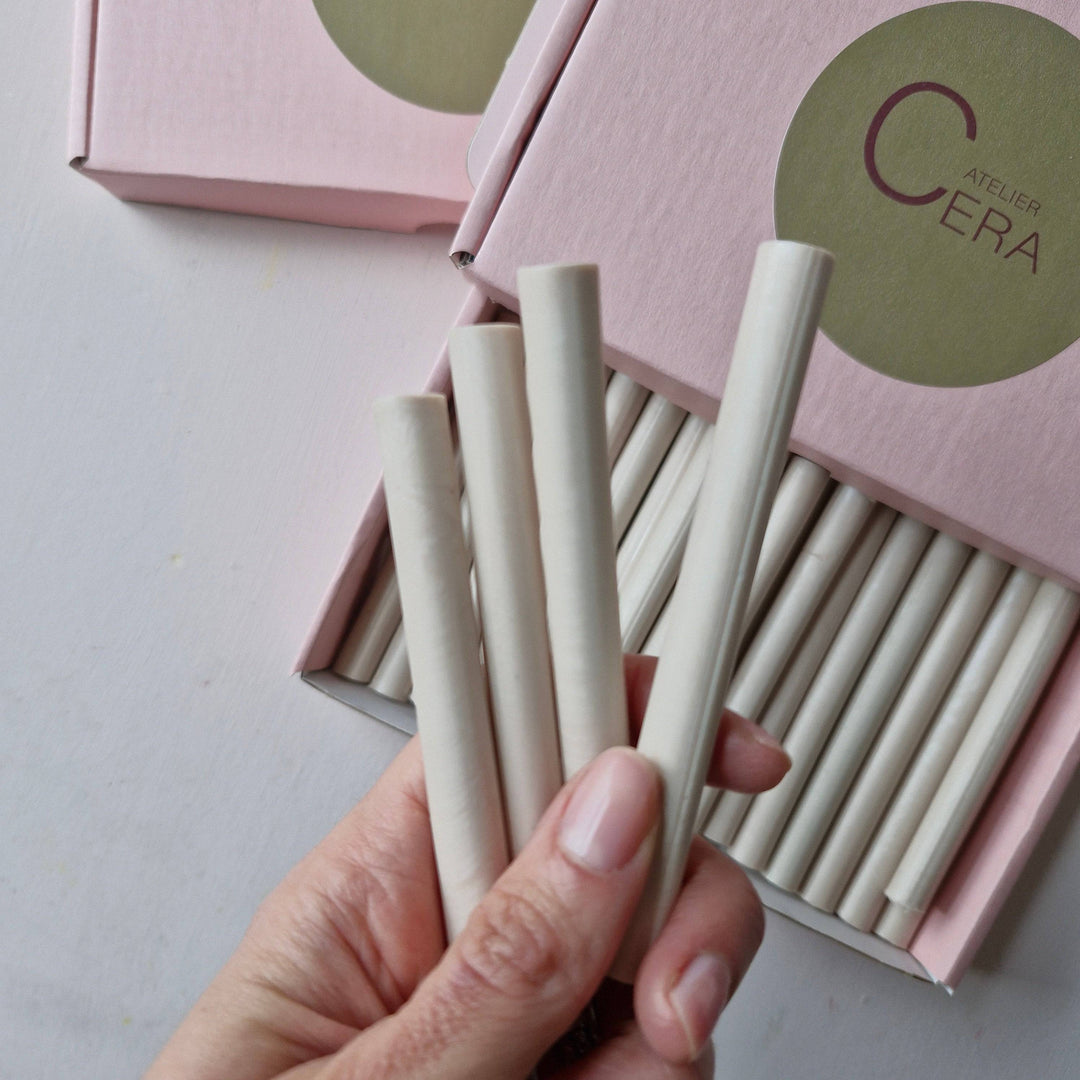 Hand holding 11mm champagne pearl sealing wax sticks with a pink box labeled 'Atelier Cera' in the background. Box contains 30 handmade sticks.