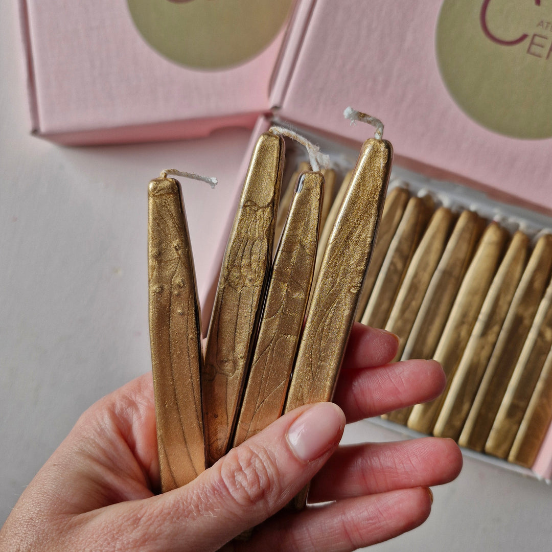 Close-up of a hand holding gold sealing wick wax sticks with a pink box in the background, highlighting their smooth metallic design.