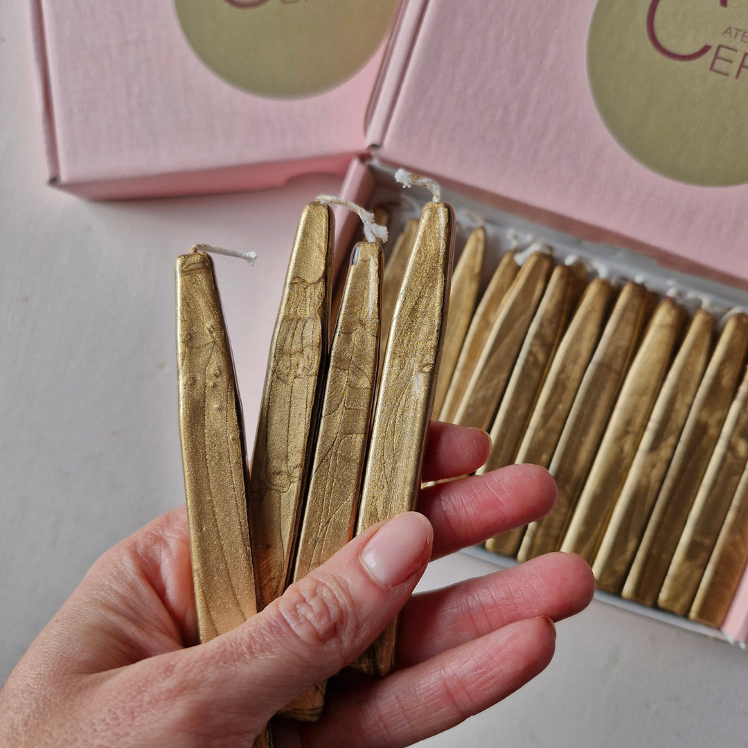 Hand holding multiple gold sealing wick wax sticks from a 26-count box, showcasing their metallic finish and detailed texture.