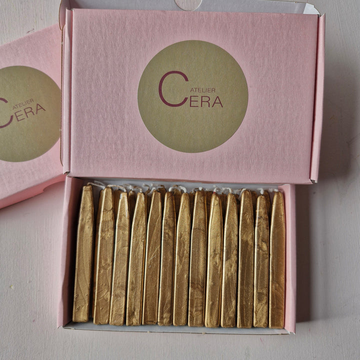 Open pink box containing 26 gold sealing wick wax sticks neatly arranged, featuring the Atelier Cera logo on the lid.