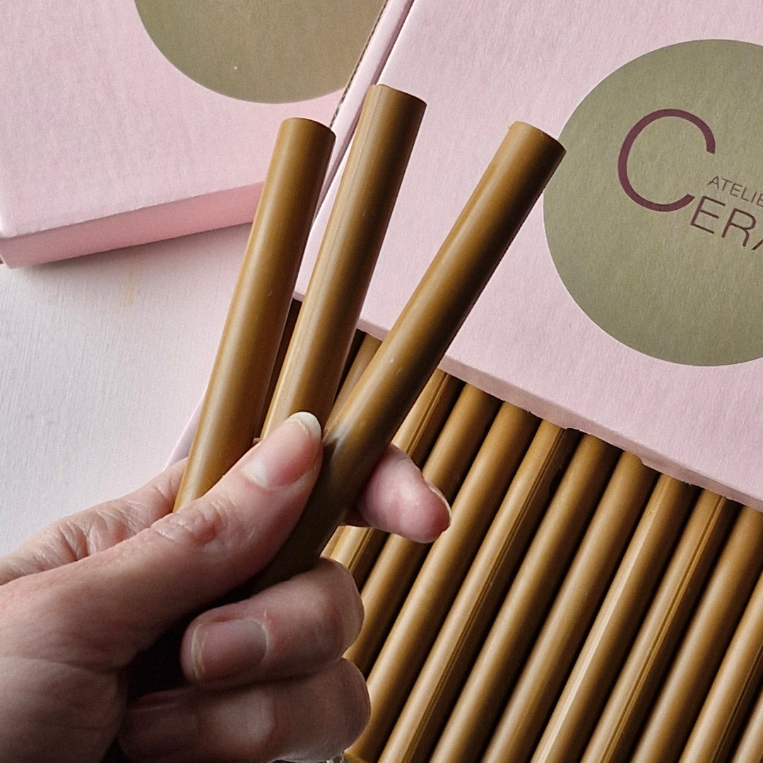 Close-up of a hand holding three toffee-colored 11mm sealing wax sticks. Pink box with logo and additional sticks visible in the background.