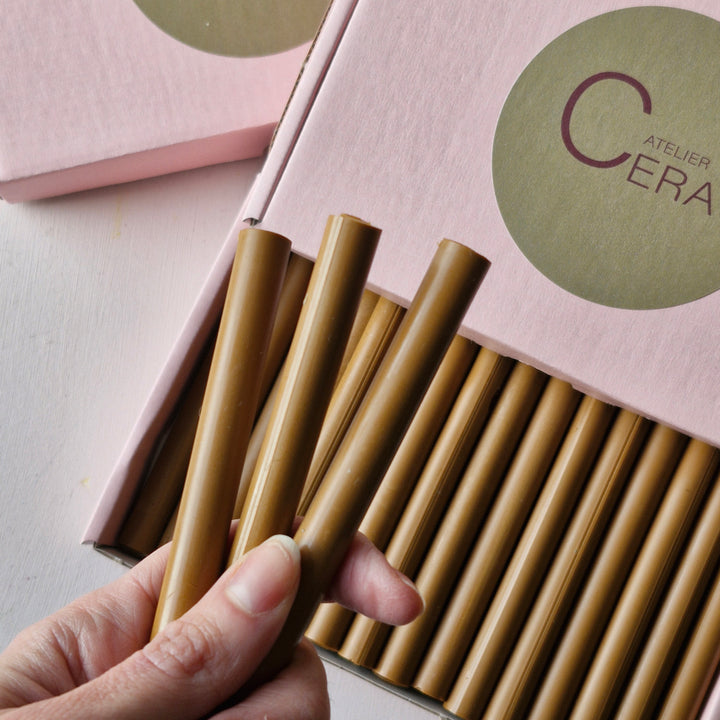 Hand holding three toffee-colored sealing wax sticks in front of an open pink box containing multiple 11mm wax sticks. Ideal for crafts and sealing.