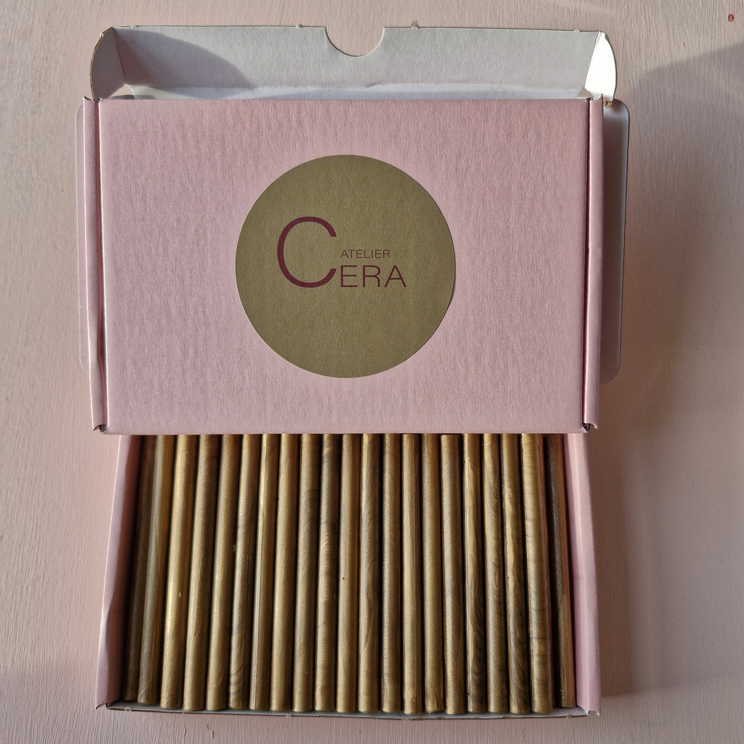 Open pink box labeled 'Atelier Cera' showcasing neatly arranged gold 7mm sealing wax sticks for crafting and sealing.