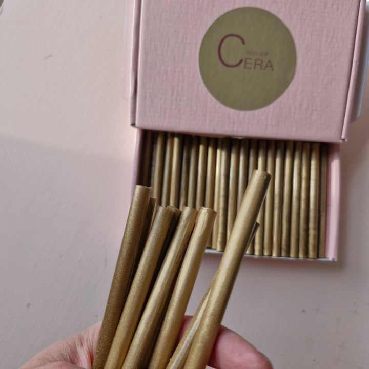 Hand holding gold 7mm sealing wax sticks, with a pink box labeled 'Atelier Cera' containing more sticks in the background.