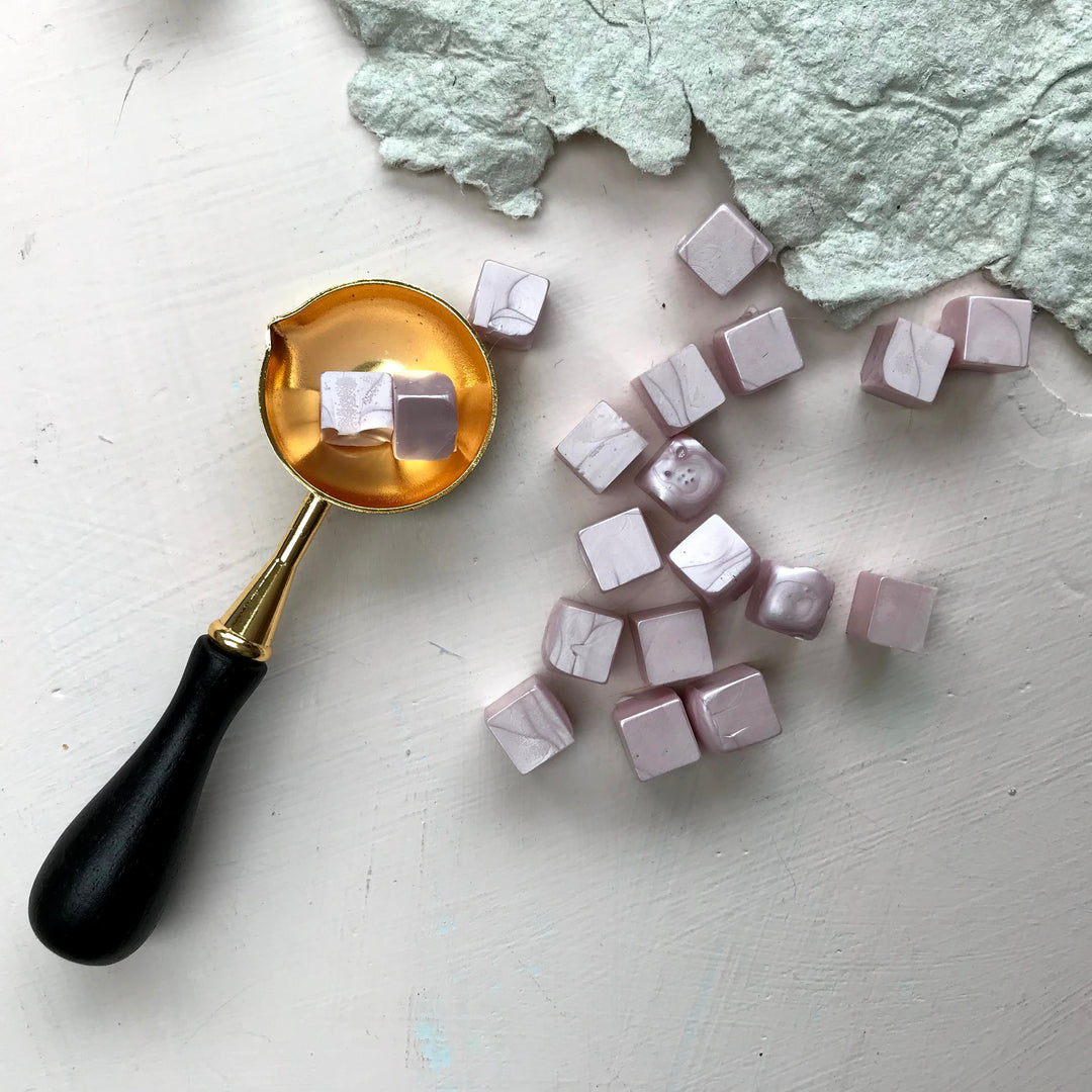 Blush-colored sealing wax beads and a gold melting spoon on a white surface, designed for creating 25mm elegant letter seals.