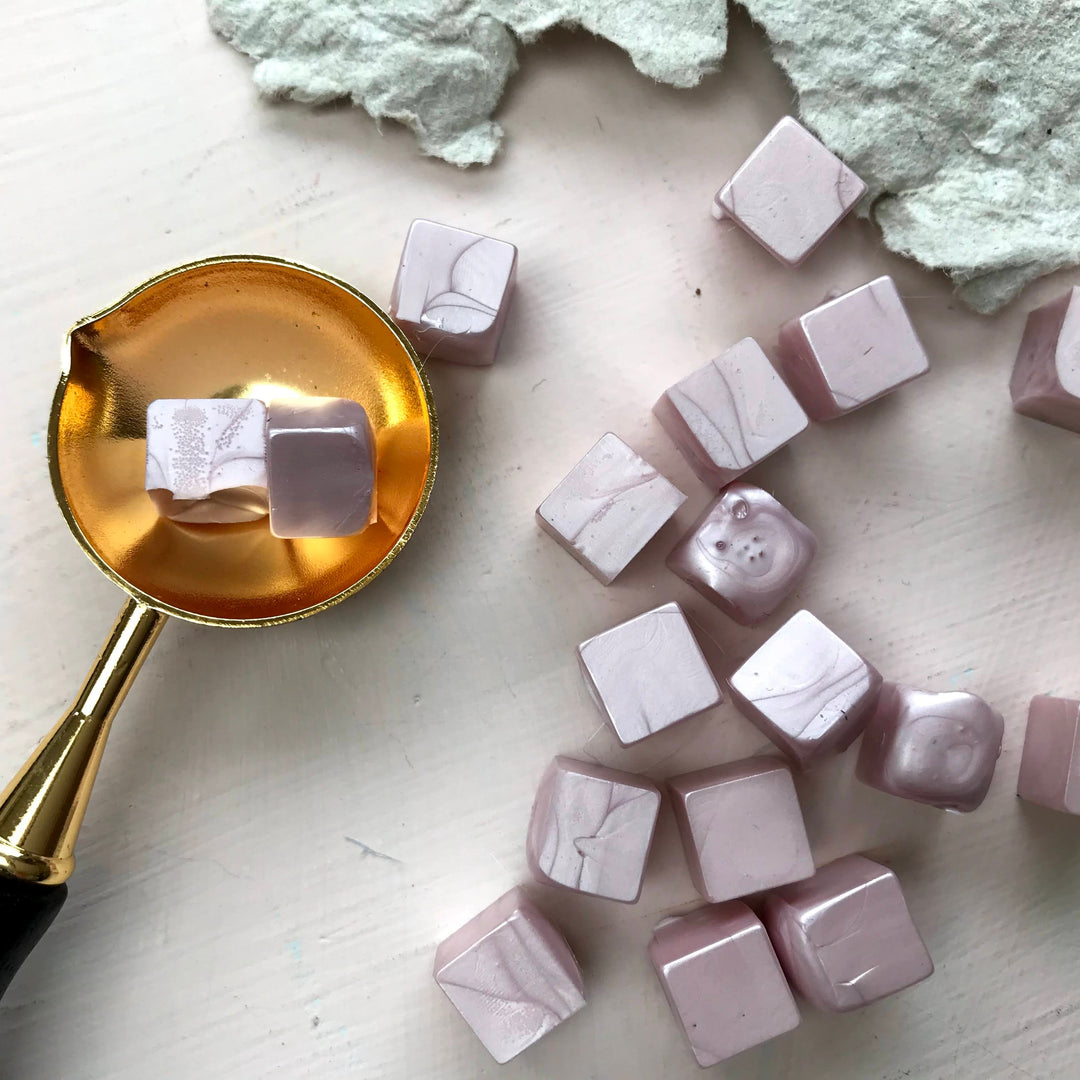 Blush sealing wax beads with a marbled finish displayed with a gold melting spoon on a textured surface. Ideal for 25mm letter seals.