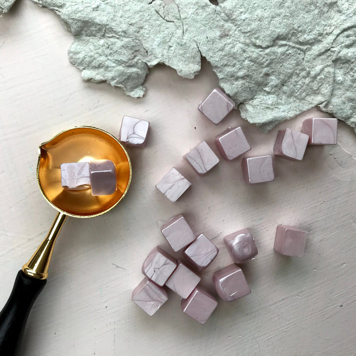 Marbled blush sealing wax beads paired with a gold melting spoon, suitable for crafting elegant 25mm seals for letters and crafts.