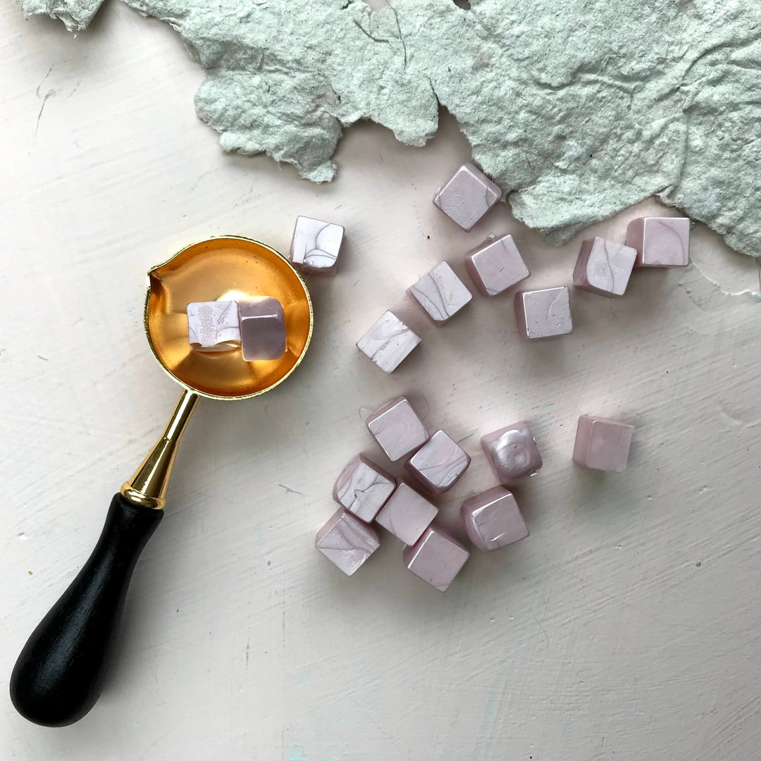 Elegant blush sealing wax beads arranged next to a gold melting spoon, perfect for crafting 25mm seals on letters and invitations.