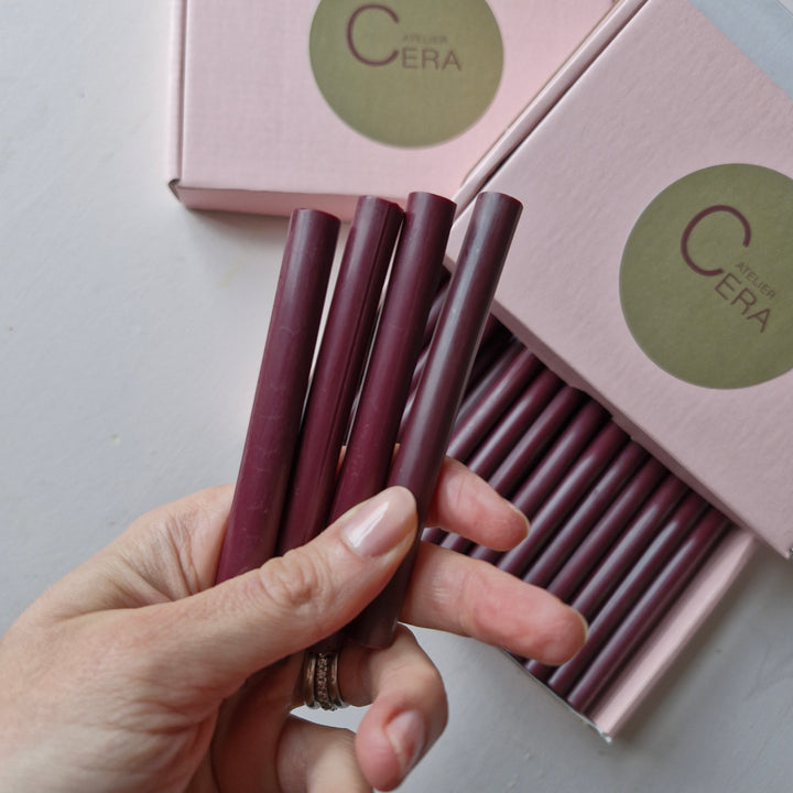 Close-up of hand holding burgundy 11mm wax sticks from a box of 30, with pink Atelier Cera packaging in the background.