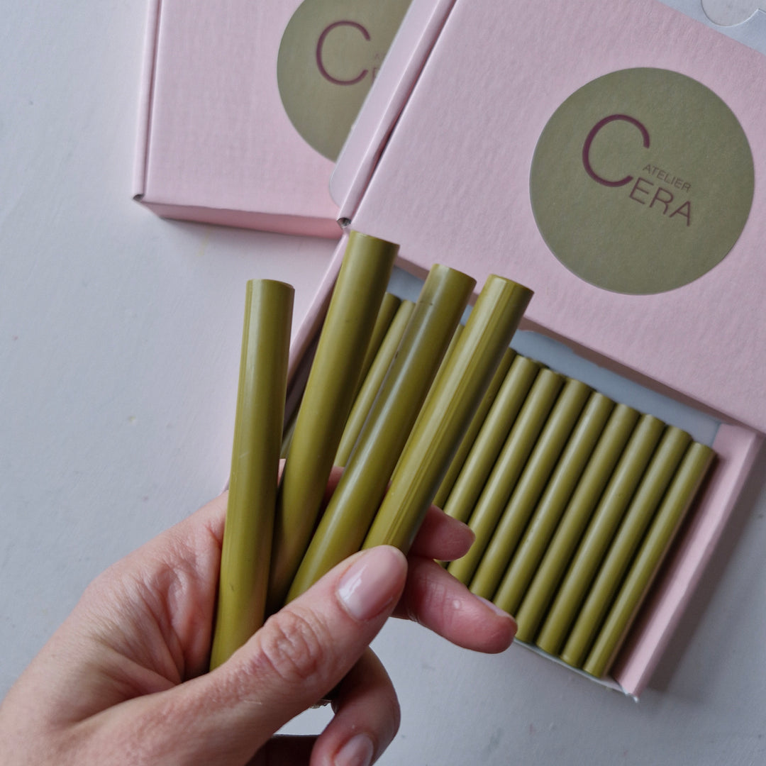 Hand holding Dijon sealing wax sticks in olive green color, with a pink box containing 11mm wax sticks in the background.