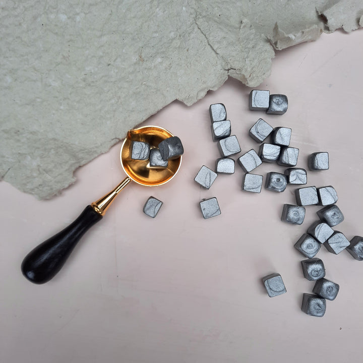 Silver sealing wax beads scattered on a light surface with a gold melting spoon and textured paper. Suitable for 25mm or 30mm seals.