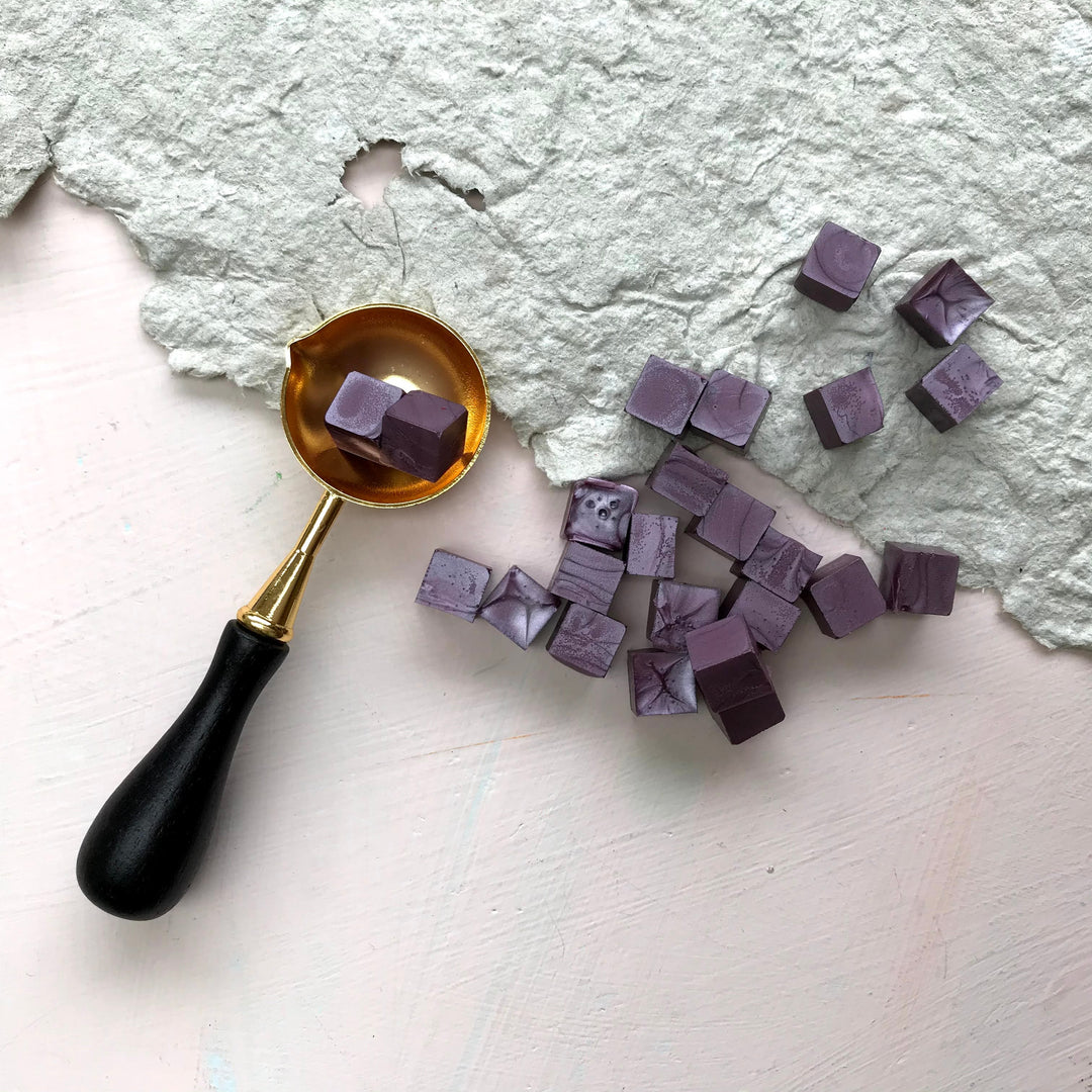 Mauve sealing wax beads scattered on textured paper with a gold melting spoon. Perfect for quick and easy wax seal crafting.