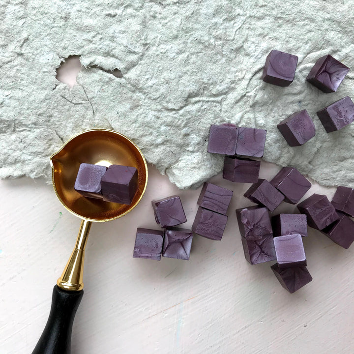 Close-up of mauve sealing wax beads and a gold melting spoon on textured paper. Designed for 25mm wax seals with fast dispatch.