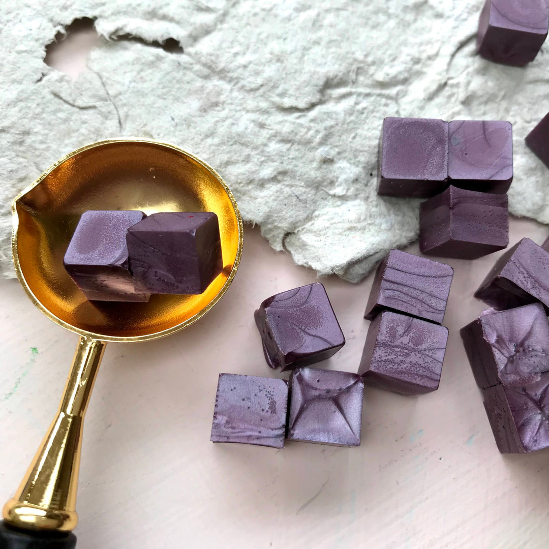 Mauve sealing wax beads displayed with a gold melting spoon on textured paper. Ideal for creating elegant 25mm wax seals.