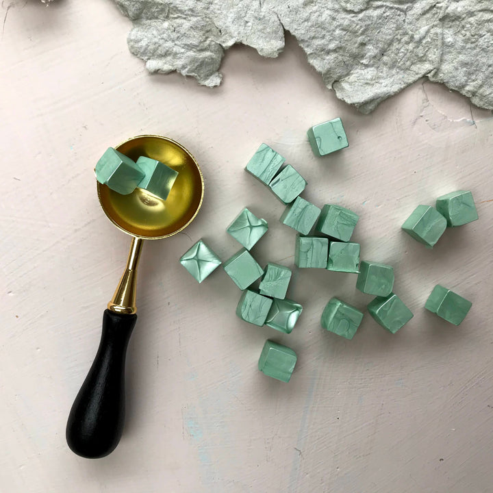 Sage green sealing wax beads and a gold wax spoon on a light textured background. High-quality wax for crafting and sealing.