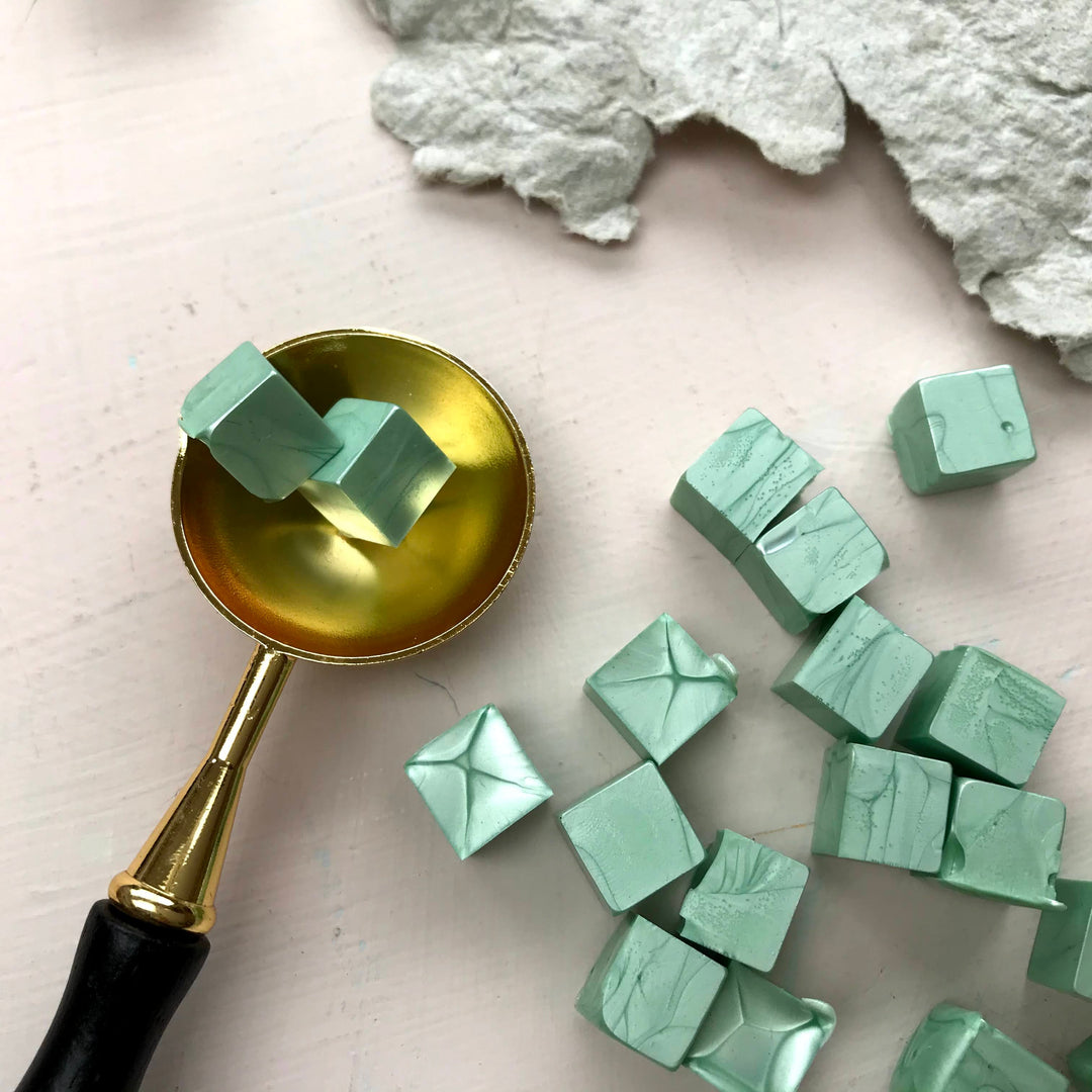 Sage green sealing wax beads with a smooth texture displayed with a gold wax spoon on a light background. Ideal for crafting elegant seals.