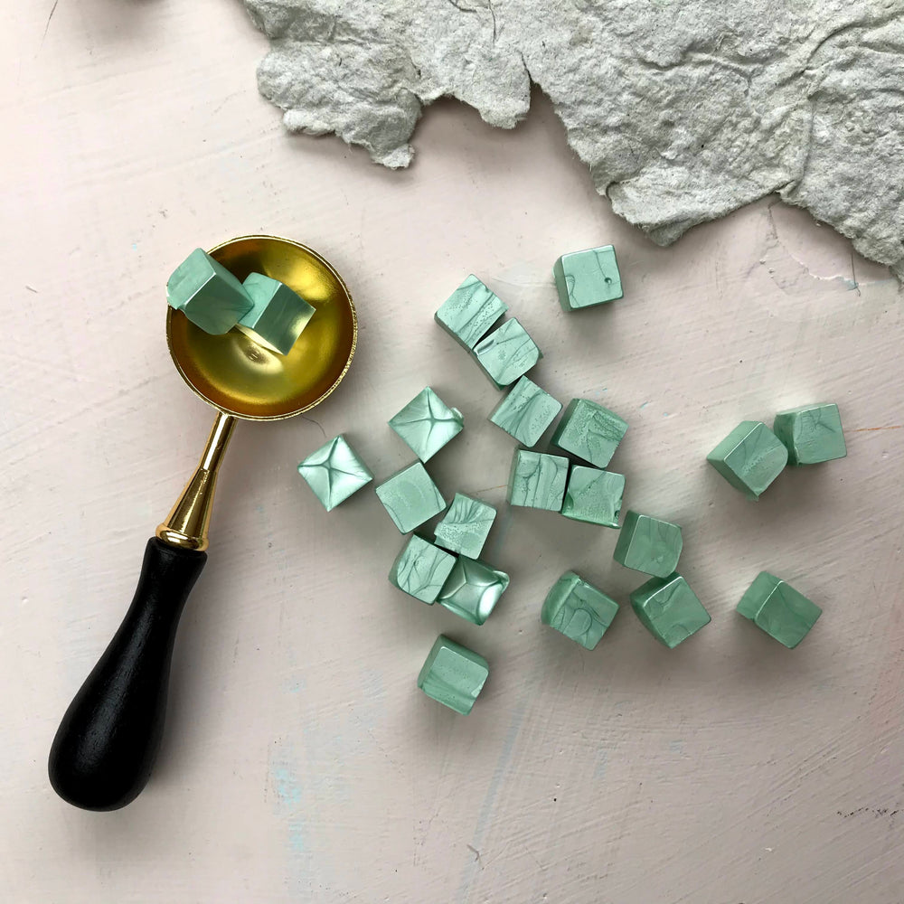 Luxurious sage green sealing wax beads arranged with a gold wax spoon on a textured surface. Perfect for creative sealing projects.